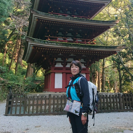 周囲の木々が生い茂り、三重塔を背景に、リュックサックを背負った西浦 里美さんが笑顔で写っている写真