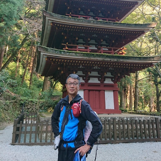 周囲の木々が生い茂り、三重塔を背景に、西浦勝也さんが腰に手をあてるポーズをしている写真