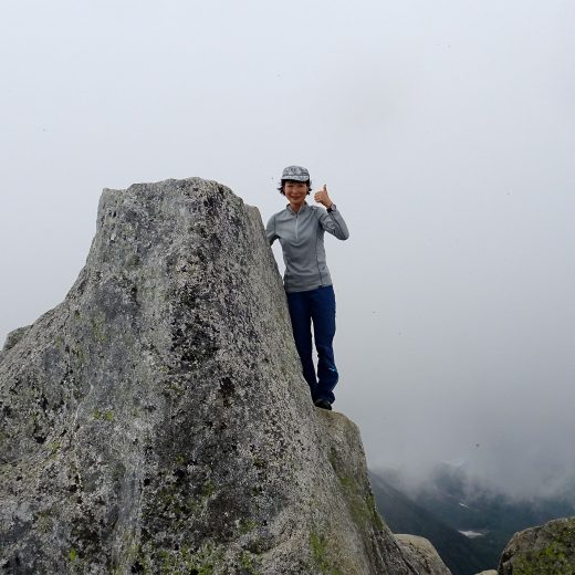 山頂の大きな岩に立ち、左手親指を上に立てたポーズをしている亀田 由香子さんの写真