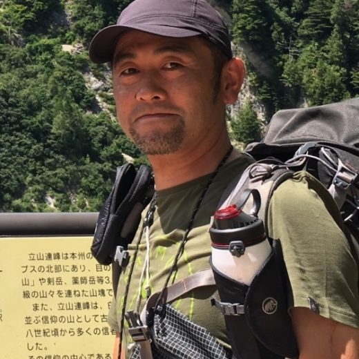 緑の山々を背景に、リュックサックを背負った野中 太郎さんをアップで写した写真