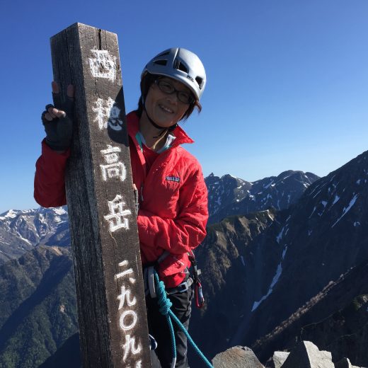 「西穂高岳2,909メートル」と書かれた独立標高点に立ち、右手でピースサインをしている清水典子さんの写真
