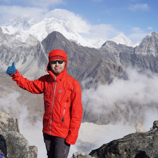 雪が積もった山頂を背景に、赤色のジャケットを着用し、右手親指を上に立てたポーズをしている武藤 直さんを写した写真