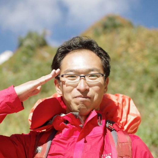 眼鏡をかけ、赤色のフード付きジャケットを着用した田村 茂樹さんが敬礼のポーズをしている写真